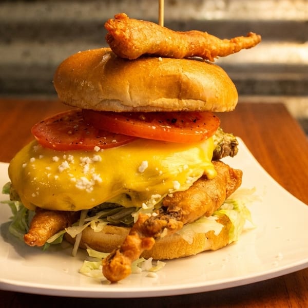 A towering burger with melted cheese, tomatoes, lettuce, and crispy fried shrimp on a white plate.