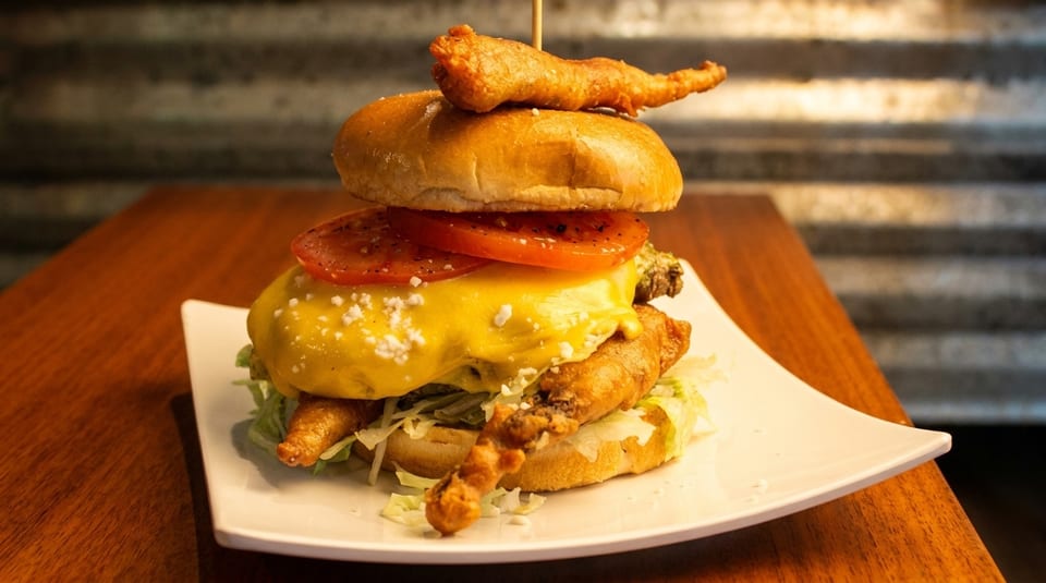 A towering burger with melted cheese, tomatoes, lettuce, and crispy fried shrimp on a white plate.