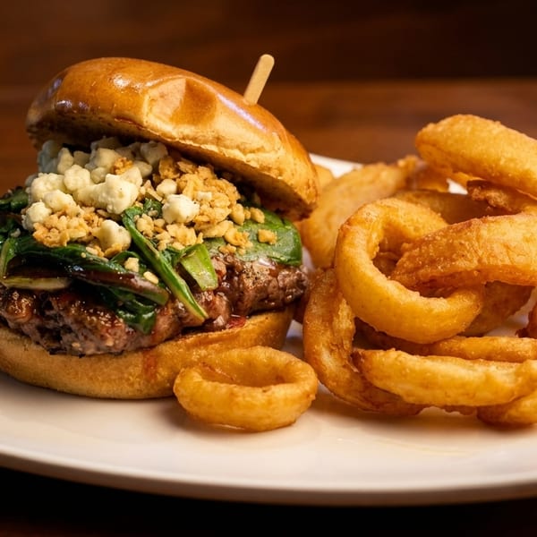 Gourmet burger with blue cheese, greens, and crispy onion rings on a white plate.