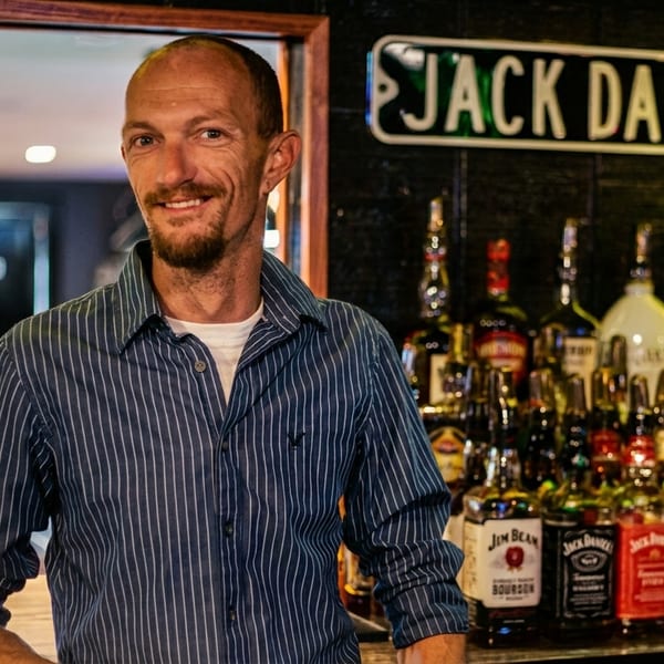Smiling bartender in a striped shirt stands behind a bar with many liquor bottles and a Jack Daniel's sign.