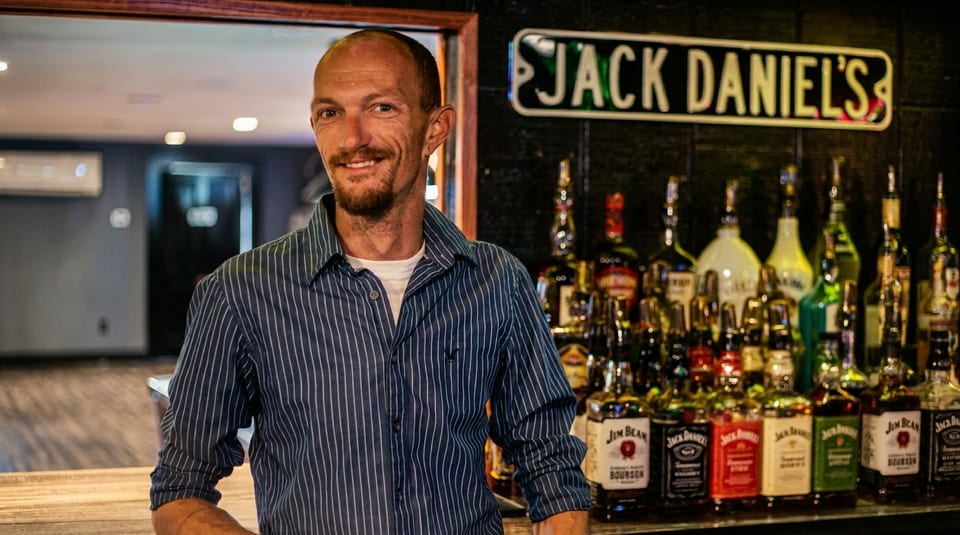 Smiling bartender in a striped shirt stands behind a bar with many liquor bottles and a Jack Daniel's sign.