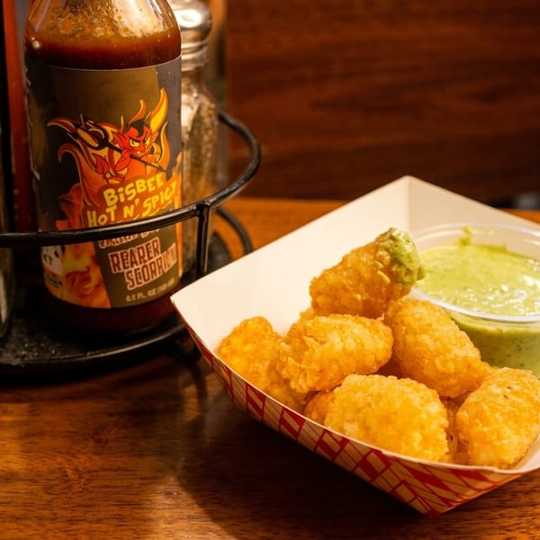 Crispy tater tots with green dipping sauce in a paper tray, next to salt and hot sauce.