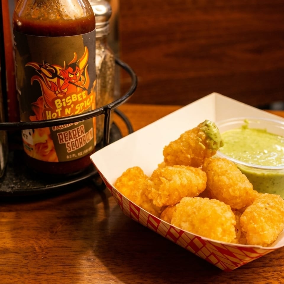 Crispy tater tots with a side of green dipping sauce, salt shaker, and hot sauce bottle.