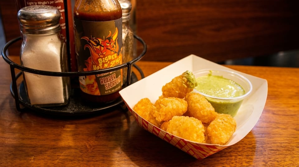 Crispy tater tots with green dipping sauce in a paper tray, next to salt and hot sauce.