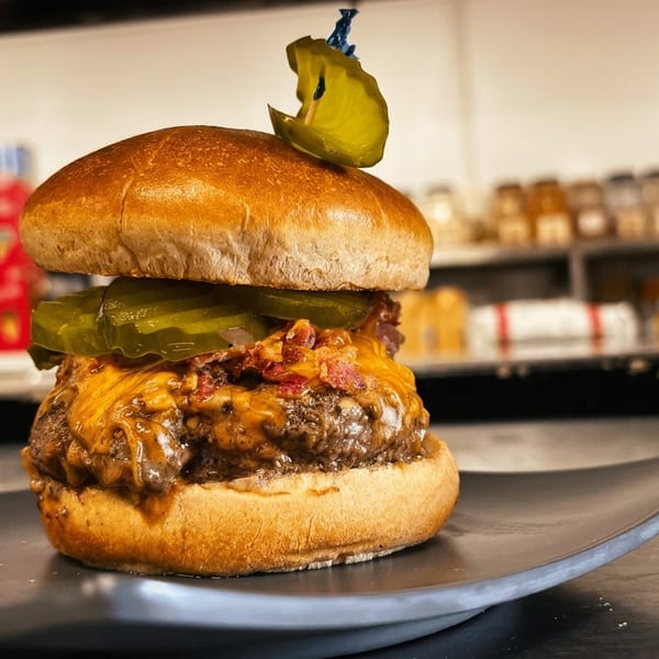 A juicy cheeseburger with bacon, pickles, and melted cheese on a dark plate in a kitchen setting.
