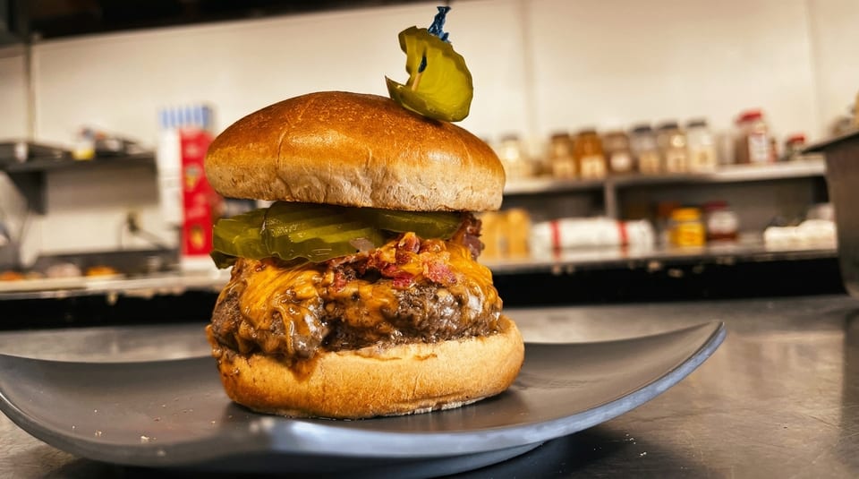 A juicy cheeseburger with bacon, pickles, and melted cheese on a dark plate in a kitchen setting.