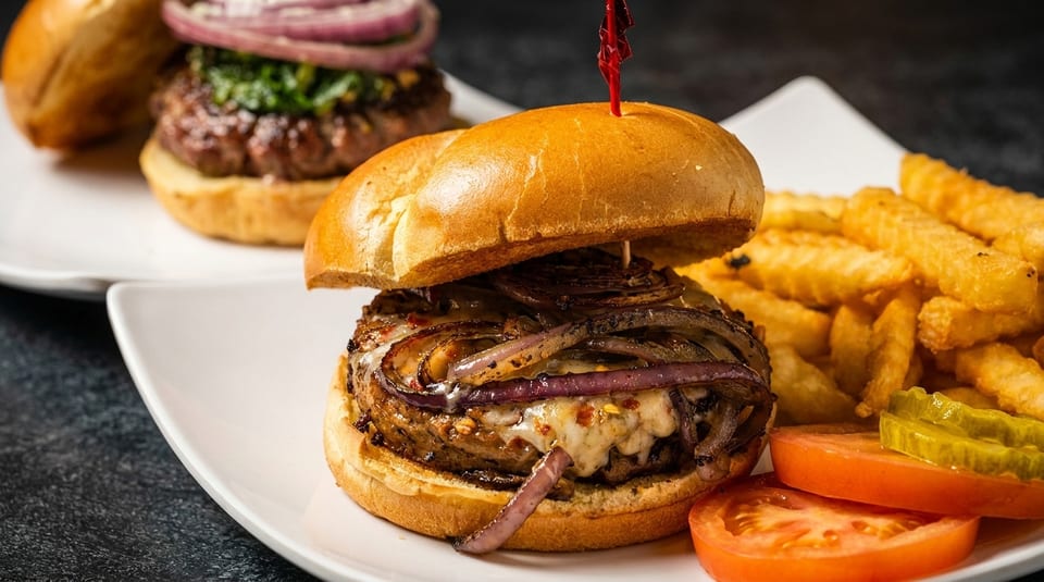 Gourmet burger with caramelized onions and melted cheese, served with crinkle-cut fries, tomato, and pickles.