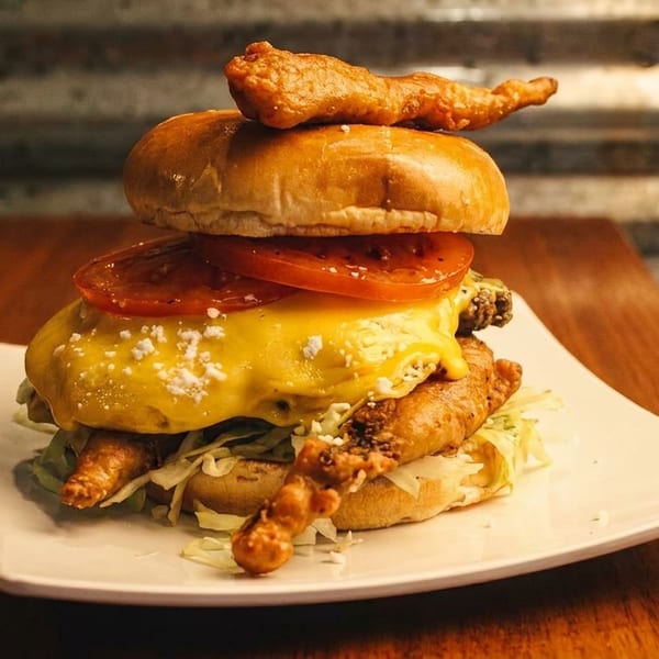 Tall burger with fried chicken, melted cheese, tomatoes, and lettuce on a white plate.