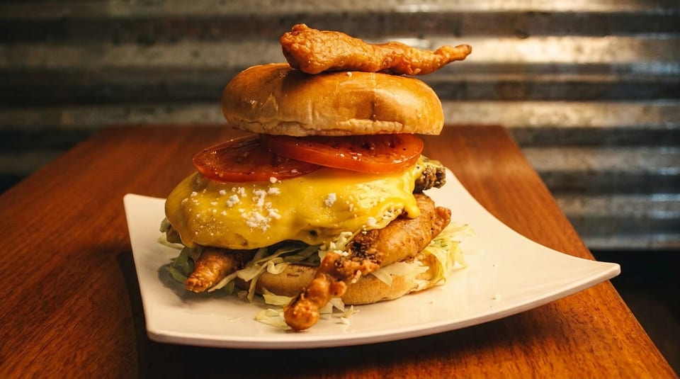 Tall burger with fried chicken, melted cheese, tomatoes, and lettuce on a white plate.